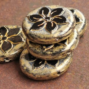 May include: A stack of antique-style, round beads with a floral design. The beads are primarily gold with black floral accents. The beads are stacked on a textured, brown surface. These beads are suitable for jewelry making.