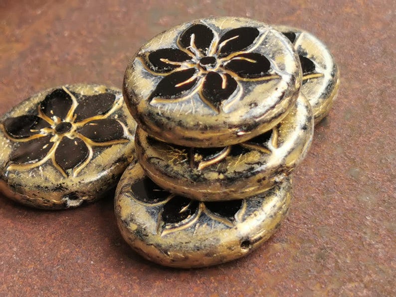 May include: A stack of antique-style, round beads with a floral design. The beads are primarily gold with black floral accents. The beads are stacked on a textured, brown surface. These beads are suitable for jewelry making.