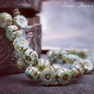 May include: A strand of pale green glass beads with a flower design. The beads are strung together to form a necklace or bracelet. The beads are on a dark brown surface.