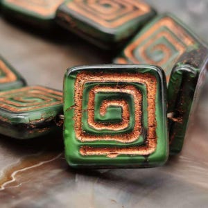 Può includere: Perle di vetro verde con un design a spirale color rame. Le perle sono di forma quadrata e hanno una finitura lucida.
