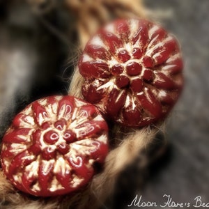 Könnte beinhalten: Zwei rot-goldene, blütenförmige Perlen. Die Perlen haben ein detailliertes Blumendesign mit einem zentralen Cluster aus kleinen Kreisen. Der Hintergrund ist verschwommen, mit einem weichen Fokus auf die Perlen. Der Text "Moon Flare's Beads" befindet sich unten rechts.