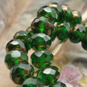 May include: Close-up of a strand of faceted, emerald green beads. The beads have a metallic gold backing, creating a shimmering effect. The beads are strung together, suggesting they are for jewelry making or crafting.