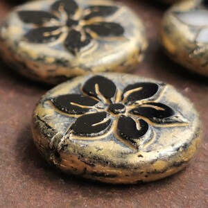 May include: Close-up of several round, decorative beads. Each bead features a black floral design on a gold-colored base. The beads have a textured, aged appearance, suggesting a vintage or handcrafted style. The background is a blurred, brown surface.