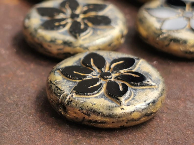 May include: Close-up of several round, decorative beads. Each bead features a black floral design on a gold-colored base. The beads have a textured, aged appearance, suggesting a vintage or handcrafted style. The background is a blurred, brown surface.