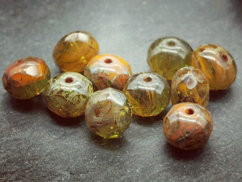 May include: A collection of ten faceted glass beads in shades of green, yellow, and orange. The beads have a mottled, marbled appearance.