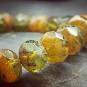 May include: A close-up of a bracelet made of faceted, green and gold glass beads. The beads are arranged in a circular pattern and have a shiny, reflective surface.