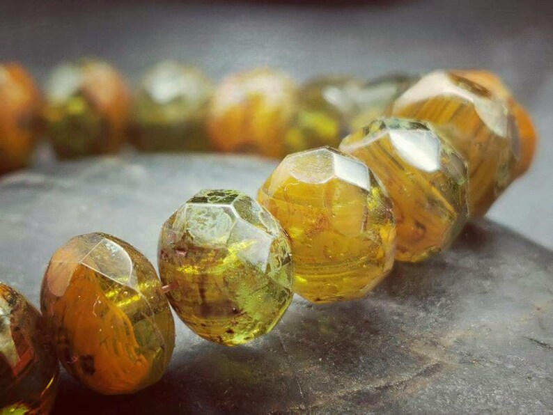 May include: A close-up of a bracelet made of faceted, green and gold glass beads. The beads are arranged in a circular pattern and have a shiny, reflective surface.