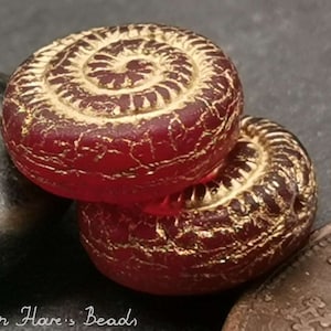 Könnte beinhalten: Zwei rote Glasperlen mit goldenen Akzenten. Die Perlen haben ein Spiraldesign und eine strukturierte Oberfläche.