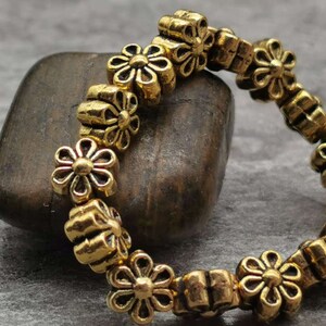 May include: A gold-toned bracelet made of small flower-shaped beads. The bracelet is lying on a brown rock.