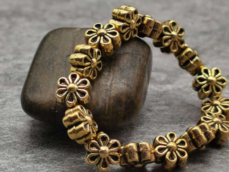 May include: A gold-toned bracelet made of small flower-shaped beads. The bracelet is lying on a brown rock.