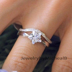 May include: A silver ring set featuring a star-shaped diamond engagement ring and a curved band with small accent stones. The rings are displayed on a finger, with a soft-focus floral background.