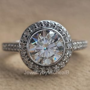 May include: A silver ring with a large, round, clear gemstone surrounded by smaller stones. The band is also set with small stones. The ring is on a neutral background. The text "JewelrybyMichealB" is at the bottom.