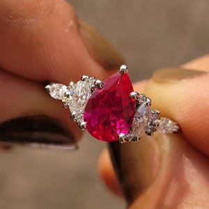May include: A silver-toned ring featuring a large, pear-shaped, vibrant red center stone. The center stone is flanked on each side by two smaller, marquise-cut clear stones set in a metallic band, creating a symmetrical and detailed jewelry piece.