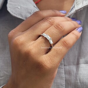 May include: A silver ring with five emerald-cut clear stones set in a curved band. The ring is on a person's finger.