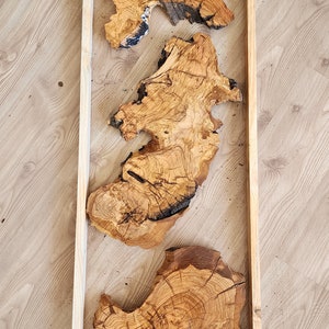 May include: Three pieces of wood with natural grain patterns are arranged in a wooden frame. The wood is a light brown colour with dark brown streaks. The frame is a light brown colour.