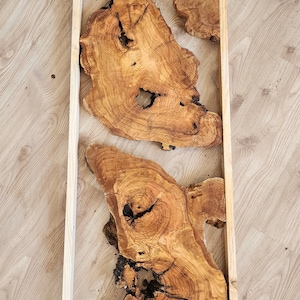 May include: A framed wall art piece featuring four natural wood slices with unique grain patterns and knots. The wood is a light brown colour and the frame is a simple, light wood.