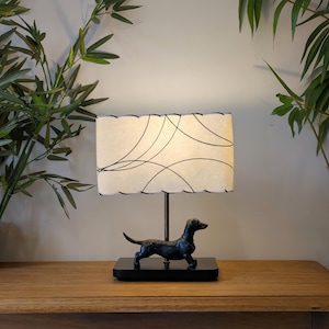 Puede incluir: Lámpara de mesa decorativa con base de perro salchicha negro. La pantalla crema tiene un diseño abstracto negro y borde festoneado. La lámpara está sobre una superficie de madera, mide unos 30 cm de alto.