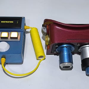 May include: A blue and yellow Ghostbusters proton pack with a digital display that reads "GHOSTBUSTERS". The pack has three yellow lights and a knob. The proton pack is connected to a yellow coiled cord.
