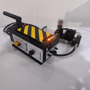 Puede incluir: Una réplica de mochila de protones de los Cazafantasmas negra y amarilla con asa, función de iluminación y varios cables y conectores.