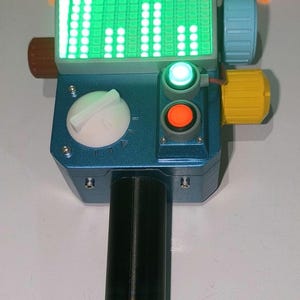 May include: A blue and black handheld device with a green LED display, a white knob, and two red and green buttons. The device has two orange antennas on top.