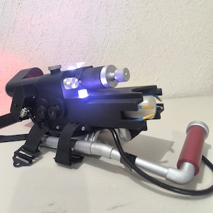 May include: A black and silver toy ghost trap with a clear plastic tube and a red and silver extension. The trap has a strap and a handle.