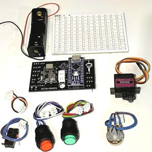 May include: An assortment of electronic components, including a black circuit board with the text "ECTO-PROPS", a battery holder, an LED array, a servo motor, push buttons, and a potentiometer. Wires and connectors are also visible.