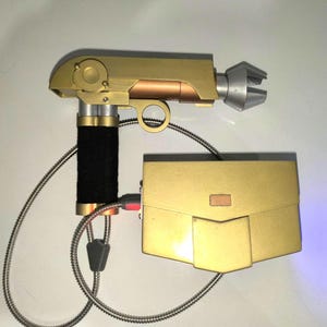 Puede incluir: Una pistola de juguete de color dorado con un aspecto futurista, con un mango negro y una punta plateada. La pistola está conectada a una caja rectangular dorada mediante un cable en espiral.