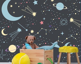 May include: A wooden toy wagon with a yellow tennis ball, a brown teddy bear, and a blue dinosaur toy. The wagon is on a light wood floor in front of a dark gray wall with a space-themed pattern of stars, planets, and constellations.