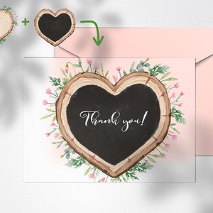 May include: A white card featuring a heart-shaped wood slice with a black chalkboard insert. The chalkboard displays the text "Thank you!" in elegant script. The wood slice is adorned with pink flowers and green foliage. The card is positioned beside a pink envelope.