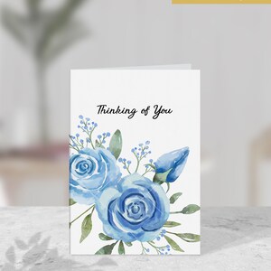 May include: A white card with the text "Thinking of You" in black script. The card is decorated with a watercolor illustration of blue roses and green leaves.