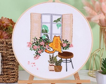 May include: A white embroidery hoop with a design of a cat sitting on a stool by a window with plants. The hoop is on a wooden easel.