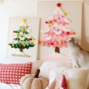 Op de afbeelding: Twee ingelijste kerstboomwanddecoraties, een groene en een roze, met pomponversieringen. Een pluche wit kussen, een bruin pompoenvormig kussen en twee dwergfiguren liggen op een wit bed.