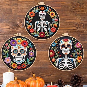 May include: Three embroidered Day of the Dead skull hoops with floral designs. The skulls are white with colorful details, surrounded by flowers in red, orange, purple, and blue. The hoops are displayed with pumpkins and candles.