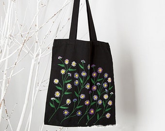 May include: Black tote bag with embroidered floral design. The design features white, yellow, and purple flowers with green stems and leaves. The bag has long black handles and is hanging against a white background.