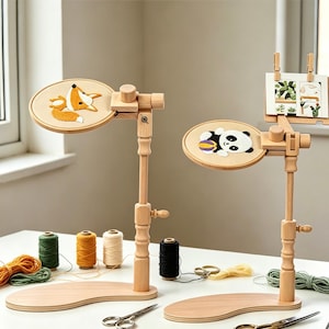 May include: Two adjustable wooden embroidery hoop stands with light wood bases. One stand holds a hoop with a fox embroidery, the other a panda embroidery and a small document clip. Spools of colored thread and metal scissors are arranged nearby.