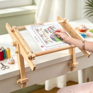 May include: A light-colored wooden embroidery frame stand with adjustable knobs and metal wing nuts, featuring a mounted cross-stitch pattern of a house and flowers on a white grid sheet, accompanied by colorful thread spools and metal scissors.