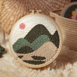 Puede incluir: Un aro de bordado circular con fondo beige y un diseño de paisaje verde, beige y blanco. El diseño presenta un sol rosa y montañas verdes.