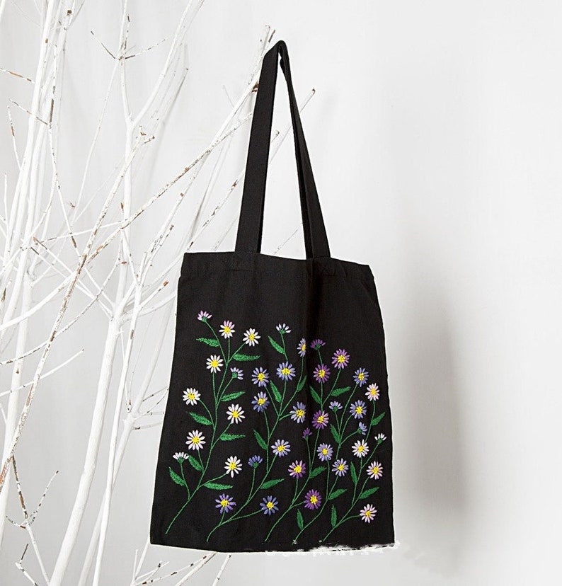 May include: Black tote bag with embroidered floral design. The design features white, yellow, and purple flowers with green stems and leaves. The bag has long black handles and is hanging against a white background.