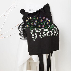May include: Black tote bag with embroidered floral design. The bag features white, yellow, and purple flowers with green stems. The bag has long black handles and is displayed on a white surface.