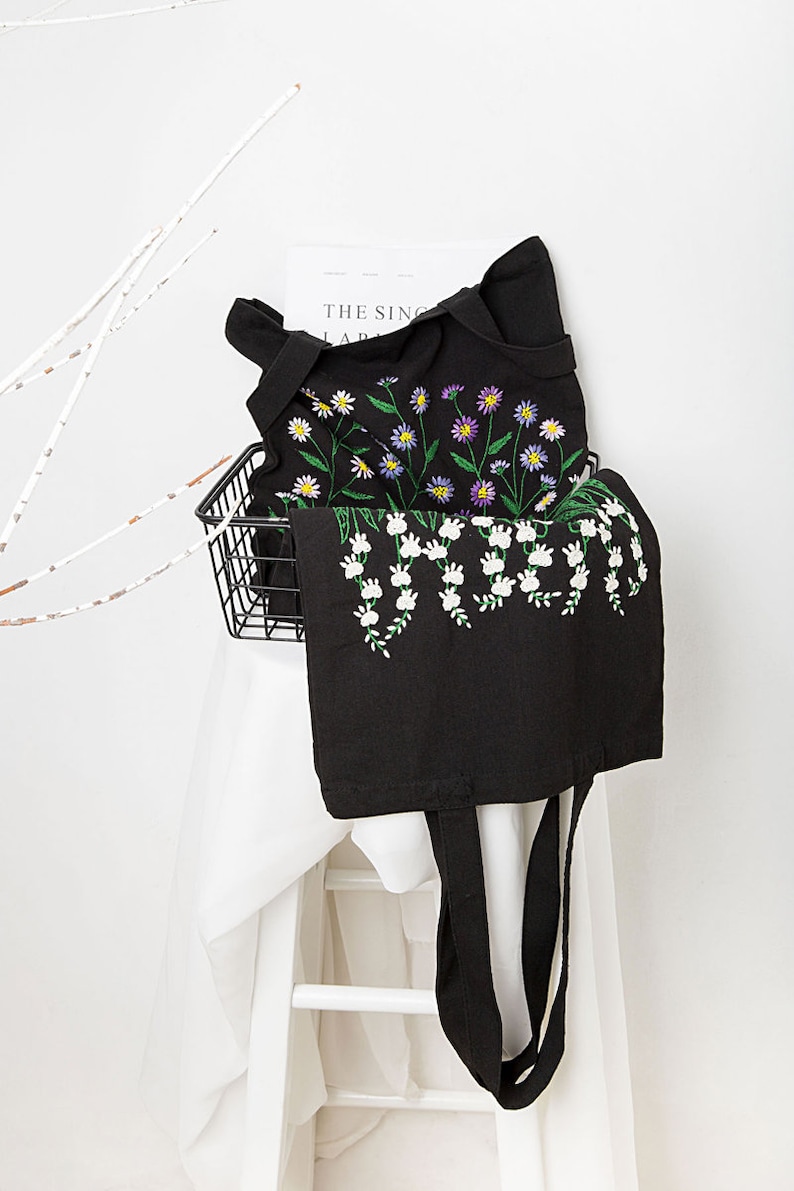 May include: Black tote bag with embroidered floral design. The bag features white, yellow, and purple flowers with green stems. The bag has long black handles and is displayed on a white surface.