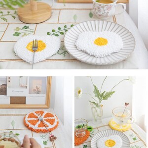 May include: Four white and yellow crocheted coasters with a variety of designs, including a fried egg, a slice of orange, and a heart. The coasters are on a white tablecloth with a floral pattern.