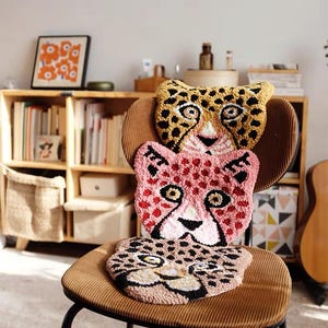 May include: Three plush animal head-shaped cushions stacked on a brown corduroy chair. The top cushion is yellow with black spots, the middle cushion is pink with red spots, and the bottom cushion is brown with black spots. All three cushions have black eyes and a white nose.
