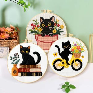 Könnte beinhalten: Drei runde Stickrahmen mit Kreuzstichmotiven von schwarzen Katzen. Eines zeigt eine Katze in einem Blumentopf, ein anderes eine Katze auf einem Bücherstapel neben einer Vase und das dritte eine Katze auf einem gelben Fahrrad mit Blumenkorb.