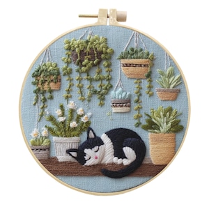 May include: A circular embroidery hoop containing a light blue fabric background with hand-stitched designs of various potted plants, hanging planters, and a sleeping black and white cat resting on a brown surface, all framed in a wooden hoop.