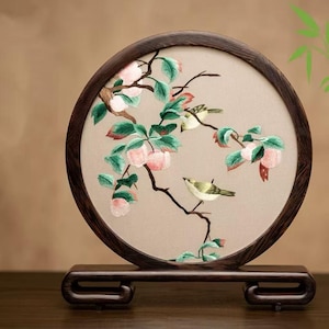May include: A circular embroidered artwork depicting a peach branch with pink fruits, green leaves, and two small birds. It's set in a dark brown wooden frame and displayed on a matching stand. The background is a neutral beige.