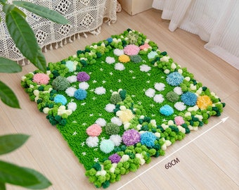 Peut inclure: Un tapis carré vert avec un motif de marguerites blanches et des pompons colorés autour des bords. Le tapis mesure 60 cm de long.