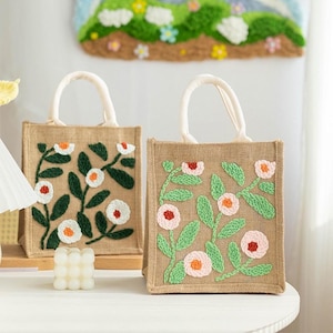 Op de afbeelding: Twee bruine jute tassen met witte handvatten. Beide tassen hebben geborduurde bloemenmotieven. De ene tas heeft witte bloemen en groene bladeren, terwijl de andere roze en witte bloemen met groene bladeren heeft.
