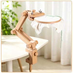 May include: A light-coloured wooden embroidery hoop stand with an adjustable articulated arm and a C-clamp base. The stand holds a circular wooden embroidery hoop containing white fabric with a single green thread stitch visible on the surface.