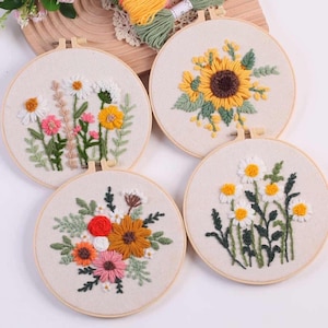 Op de afbeelding: Vier ronde borduurringen met bloemmotieven. Eén heeft een zonnebloem, een ander madeliefjes en twee andere met gemengde bloemen in roze, oranje en geel. De ringen staan tegen een neutrale stoffen achtergrond.