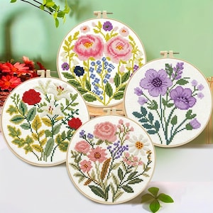 May include: Four circular wooden embroidery hoops containing cross-stitch patterns of various flowers. One hoop features pink peonies, another displays red and white lilies, a third shows purple blossoms, and the fourth depicts mixed pink and white flowers.